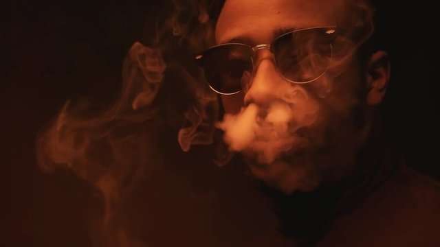Close up portrait shot with red lighting: defiant black man in sunglasses vaping and exhaling smoke from mouth and nose while looking at camera in dark studio