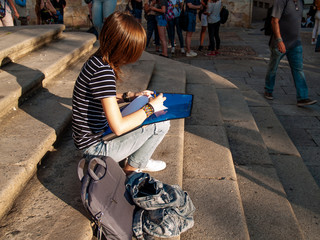 A teenage woman writing with a pen in a folio on a folder sitting in the stairs on a university...
