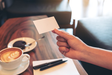 Obraz premium A hand holding white empty business card with notebook and coffee cup on wooden table