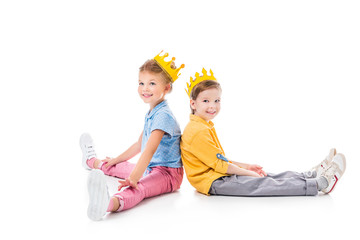 smiling friends in yellow paper crowns sitting back to back, isolated on white
