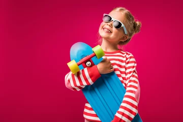 Schilderijen op glas happy stylish child in sunglasses posing with skateboard isolated on red © LIGHTFIELD STUDIOS