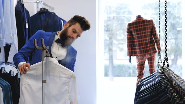 Stylish bearded seller smoothes a shirt with clothing steamer in menswear store.