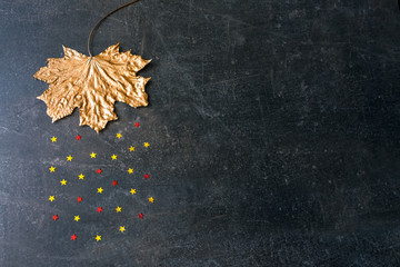 Magic gold metallic copper maple leaf with rain from the golden and red small stars. Autumn background, mockup with painted metallic fall dry leaf and star rain on dark