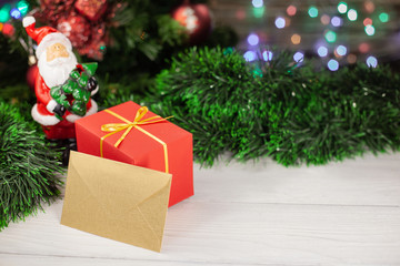 Christmas gift in red packing with a gold envelope next to a Santa Claus figure next to a Christmas tree and red Christmas balls with a ball on the background of a colored garland