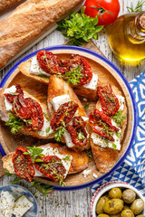 Bruschetta with feta cheese, dried tomatoes, olive oil and fresh aromatic herbs, on a plate on a wooden table.