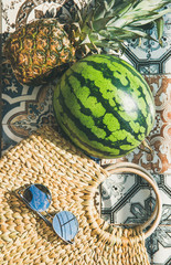 Summer lifestyle background. Flat-lay of summer fruit pineapple and watermelon, straw bag and sunglasses over colorful moroccan tile floor, top view, vertical
