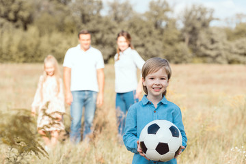 happy little kid holding ball while his family standing blurred on background in field
