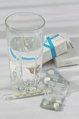 Таблетки, термометр и стакан воды на белом фоне.