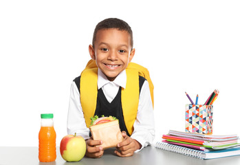 African-American schoolboy with healthy food and backpack sitting at table on white background