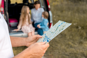 cropped shot of man holding map while his family sitting in car trunk in field