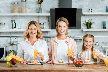 beautiful three generations of women with pancakes and orange juice for breakfast at home looking at camera