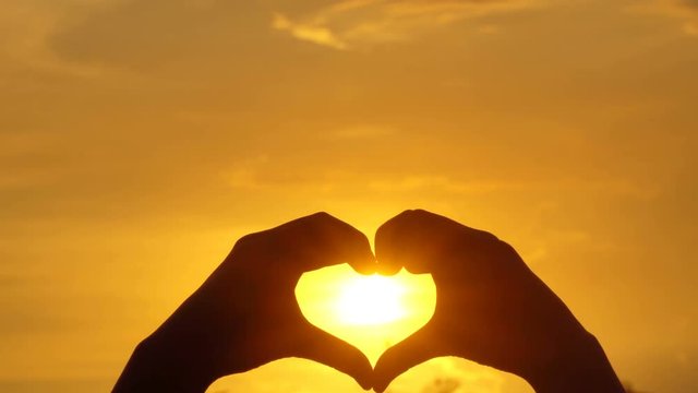 Woman shapes heart with hands over sun on sunset
