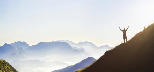 Tuinposter Man on peak of mountain. Emotional scene. Young man with backpack standing with raised hands on top of a mountain and enjoying mountain view. Hiker on the mountain top. Sport and active life concept. © bilanol