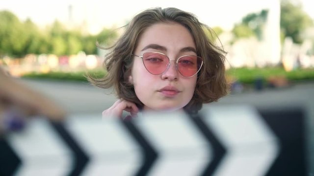 Cute young actress with short wavy fair hair in white t shirt and pink sunglasses smiling to camera and touching her hair after clapperboard being used. Advetisement. Slider slow motion portrait shot