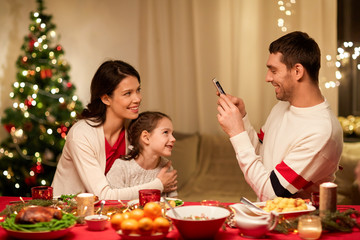 holidays, family and celebration concept - happy mother, father and little daughter having christmas dinner and taking picture by smartphone at home