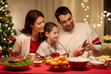 holidays, family and technology concept - happy mother, father and little daughter with smartphone having christmas dinner at home