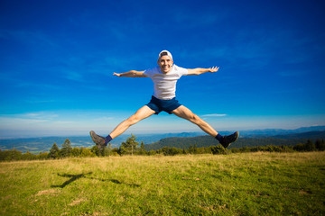 Summer hiking in mountains. Young tourist man in cap with hands up on top of mountains admires nature. Travel concept