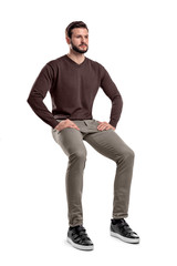 An isolated bearded man in casual wear sits on a white background with hands on his thighs.