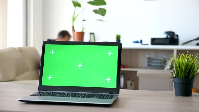 Laptop on a desk in the living room with a isolated green screen and marks for tracking. Man walks in the room and sits on the couch in the background
