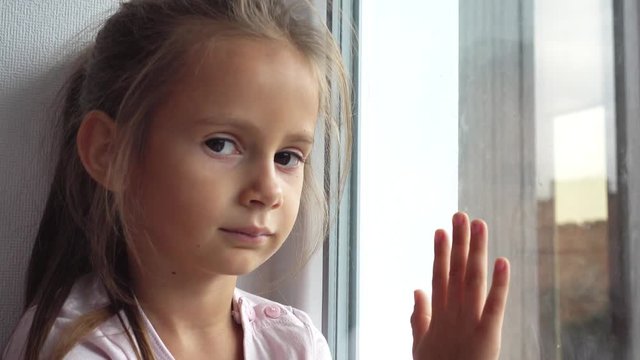 Portrait of a small sad girl by the window. Sad child looks out the window, video 4k. 
