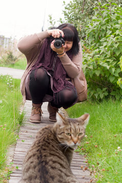 young brunette woman with camera, woman taking photo of grey striped cat outside in the park
