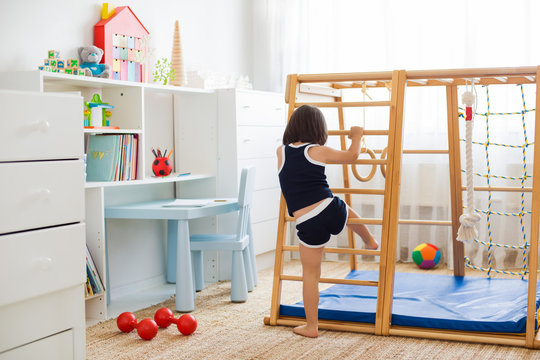 Little Girl Performs Gymnastic Exercises On A Wooden Home Sports Complex Stairs, Rings And Rope. Children's Sports Exercises. Physical Education Of Children At Home.