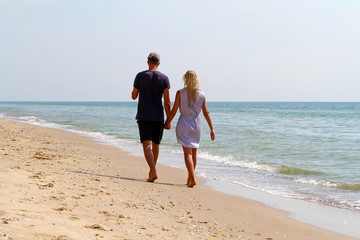 A young man and a girl with wet blond hair walk barefoot along the sandy beach from behind. A loving couple are walking along the seashore leaving the photographer ahead