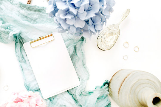 Home office desk with clipboard with copy space, turquoise blanket, colorful pastel hydrangea flower bouquet, woman fashion accessories on white background. Flat lay, top view workspace mockup.