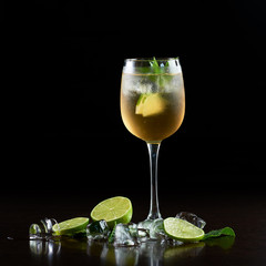 Tall crystal cocktail glass with cold refreshing juicy drink, slices of lime, fresh green mint leaves and transparent ice cubes on black background. Simplicity and elegance. Picture aspect ratio 1:1