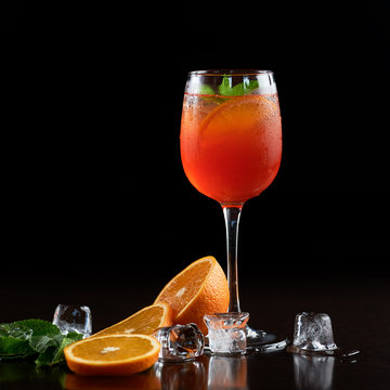 Tall crystal cocktail glass with cold summer refreshing juicy drink, slices of orange, fresh green mint leaves and transparent ice cubes on deep black background. Picture aspect ratio 1:1