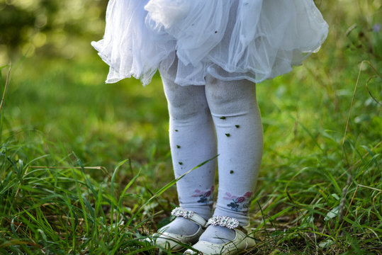 The Legs Of A Little Girl In A White Dress And Pantyhose On The Grass In The Forest Are Stained In The Ground, Grass And Thorns, Old Torn Shoes