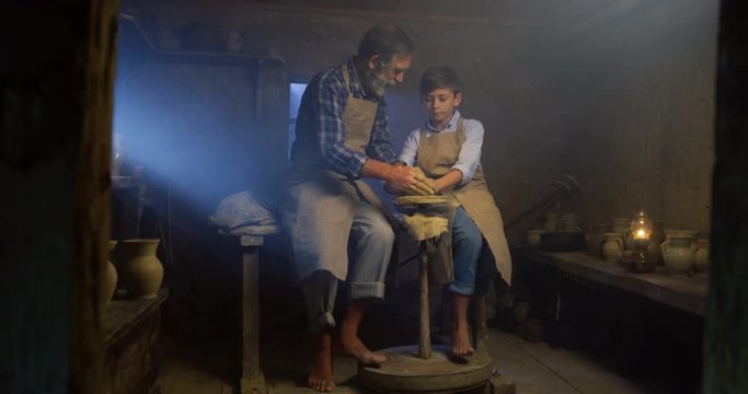 Old Caucasian man and little boy sitting in the manufactory and doing pottery thing from a clay.