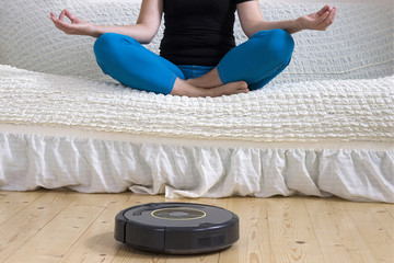 Woman sitting in lotus pose doing yoga at home while autonomous vacuum cleaner cleans the floor....