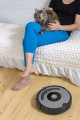 Woman carressing furry cat on the sofa near working robotic vacuum cleaner. Pet friendly modern technology for housekeeping