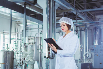A young beautiful girl in white overalls makes notes in a tablet on the background of equipment of a food processing plant. Quality control in production