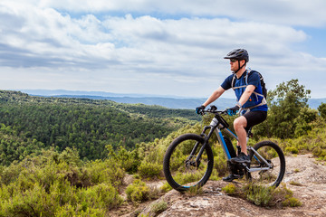 Mountain biker wearing sunglasses looking into distance on top of hill, holding one foot on the pedal