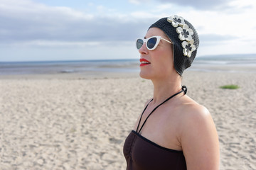 woman standing on the beach with a swimming costume on and 1950's swim hat and sunglasses