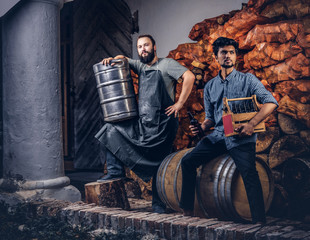Bearded brewer in apron holds barrel and his friend sitting on a wooden barrel with craft beer at brewery factory.