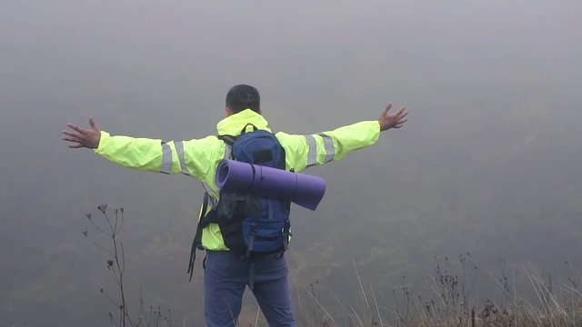 Man tourist with backpack stands on the edge of the mountain in the fog waving his hands,slow mo