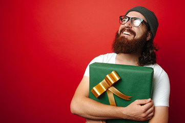 Portrait of happy cheerful bearded hipster man holding a big gift for Christmas/ New Year over red...