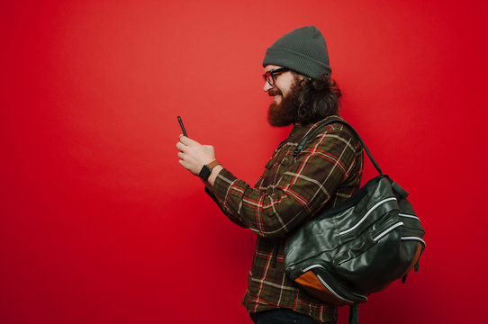 Portrait of happy bearded hipster man using smartphone over red background and wearing leather backpack. Student using phone