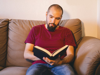 Young man with beard sitting on brown couch reading a black book.