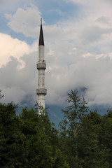 Džamija Malezija – Mosque of Malaysia – located in Turbe, Bosnia and Herzegovina. An errected minaret above trees and bushes located at the foot of a mountain with the sky full of clouds.
