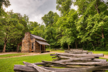 An old log and stone cabin in the green woods with an old wooden fence in the foreground and a grey...