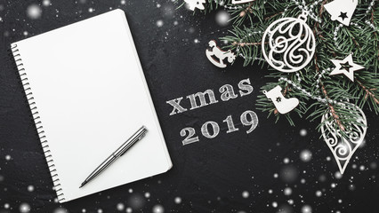 Notebook for christmas notes 2019 on black stone background. Fir tree decorated with wooden toys. Top view. Space for text. Snow effect
