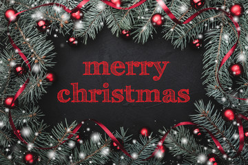 Black stone background with a frame of fir branches decorated with balls and red ribbon. Top view. With Merry Christmas text