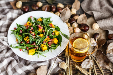 Food flat lay. Top view. Salad with mango, arugula, pomegranate seeds lying on white plate. Next lying pumpkin, cup of black tea with lemon, dry flowers and leaves, physalis, chestnuts, sweater