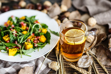 Salad with mango, arugula, pomegranate seeds lying on white plate. Next lying pumpkin, cup of black tea with lemon, dry flowers and leaves, physalis, chestnuts, sweater