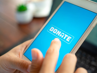 Close-up Finger pressing donate icon button on blue screen on white mobile phone with black laptop...