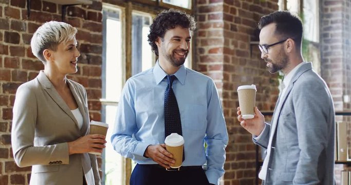 Cheerful male and female company employees standing with coffe at the break and having a talk. Indoors.